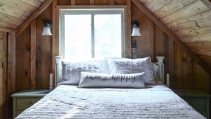 Ein Schlafzimmer mit schrägen Wänden, einem großen Fenster, einem Bett mit weißer Bettwäsche und zwei Wandleuchten.