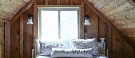 A bedroom with a sloped ceiling, a large window, a bed with white bedding, and two wall-mounted lamps.
