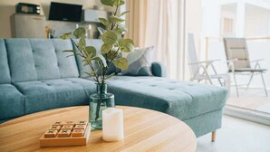Ein Wohnzimmer mit einem blauen Sofa, einem Couchtisch aus Holz mit einem Spiel und einer Kerze sowie einer Pflanze in einer Glasvase.