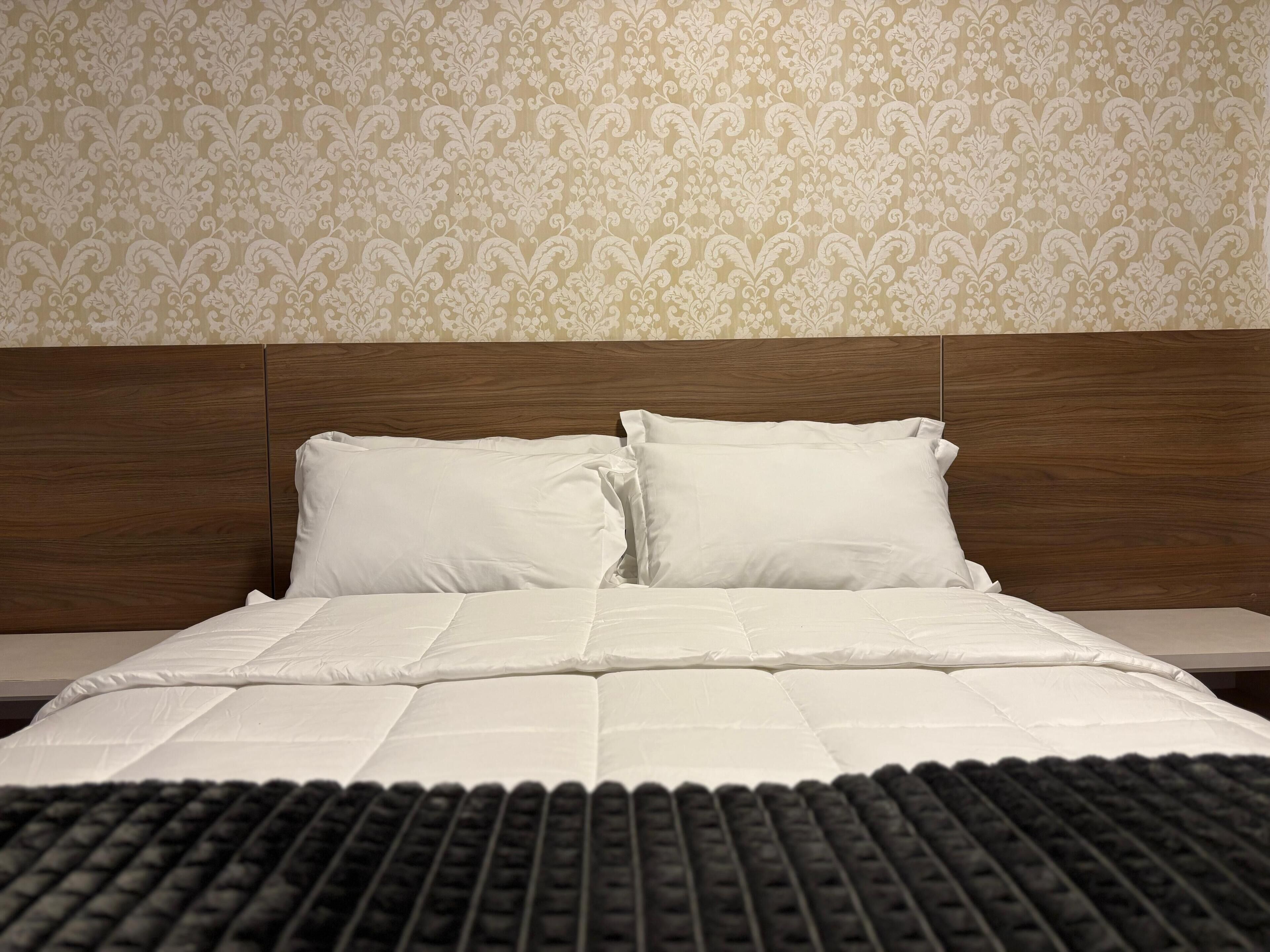 Una cama bien hecha con ropa de cama y almohadas blancas, un cabecero de madera y un fondo de papel tapiz estampado.