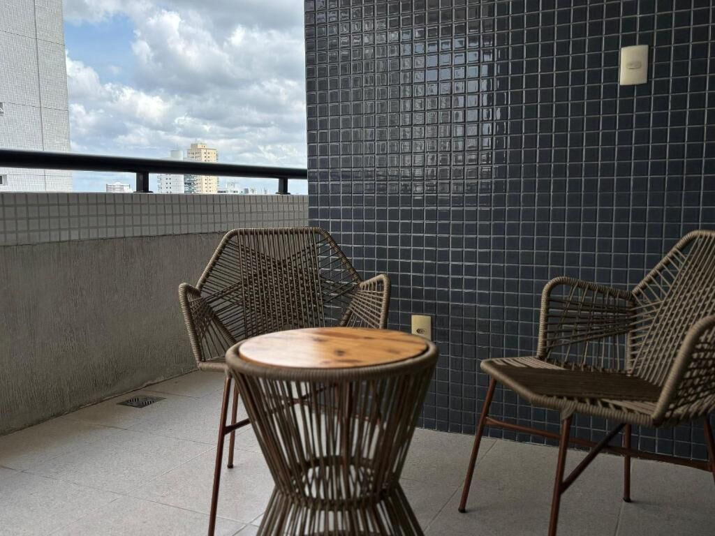 A balcony with wicker chairs and a small round table, a tiled wall, and a view of buildings under a cloudy sky.