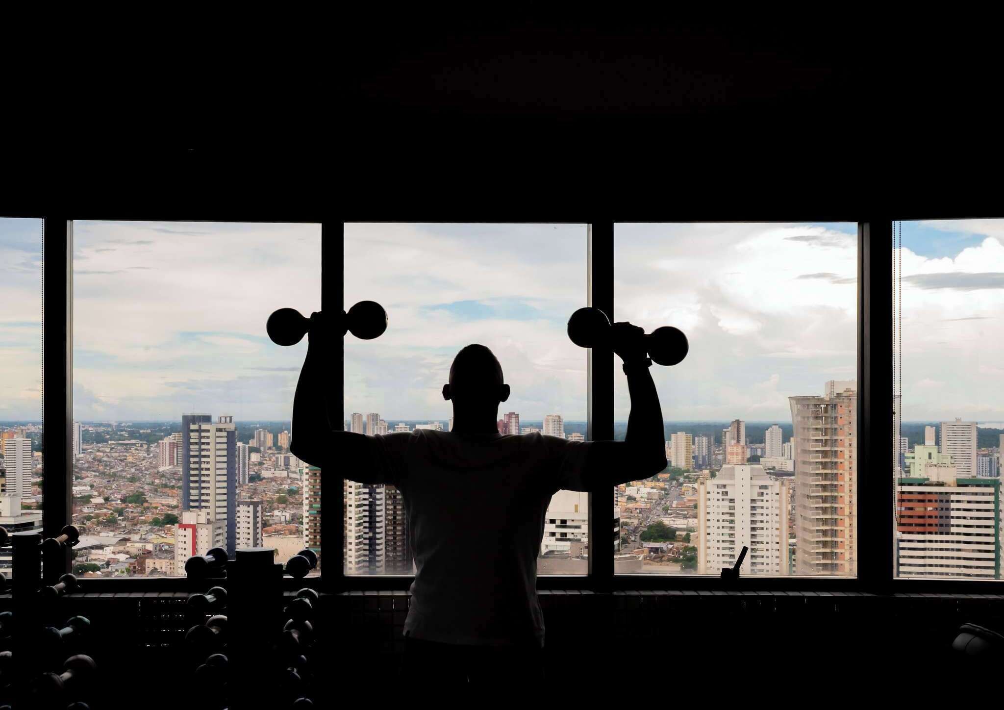 Une personne fait de l’exercice avec des haltères dans une pièce dotée de grandes fenêtres donnant sur un paysage urbain.