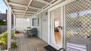 Un espace extérieur abrité, doté d’une terrasse en bois, d’une table et de chaises, et d’une porte donnant accès à un espace intérieur.
