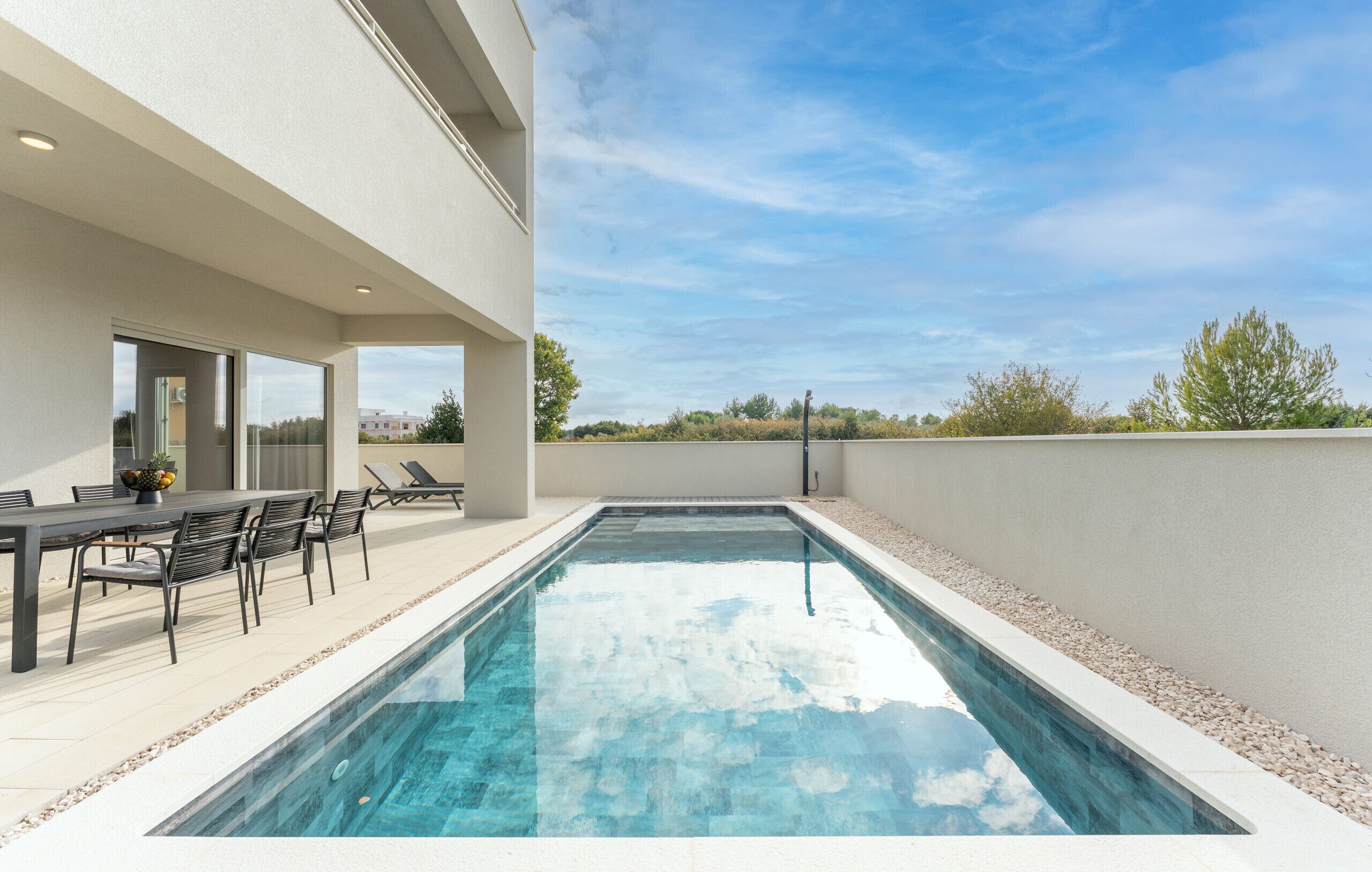 A modern outdoor pool area with a dining table and chairs, surrounded by a low wall and a clear sky.