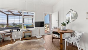 A room with a wooden floor, white walls, a large window with blinds, a television, a round table with chairs, a mirror, and a view of the sea. - Moon Gate at Culburra - Pet Friendly Absolute Beachfront (Culburra Beach)