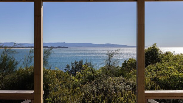 A view through a wooden-framed window overlooking a body of water with distant landforms, surrounded by dense greenery. - Moon Gate at Culburra - Pet Friendly Absolute Beachfront (Culburra Beach)