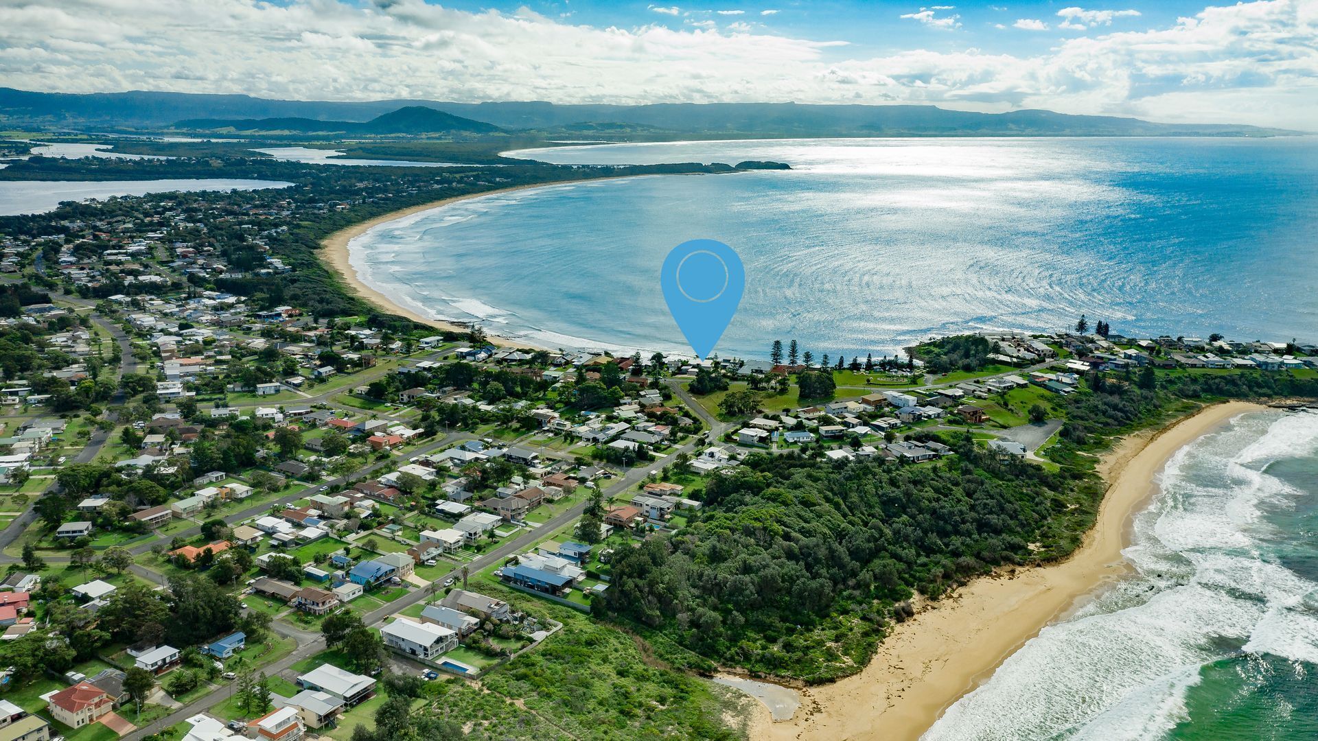 An aerial view of a coastal town with a beach, houses, and a dense forest area.