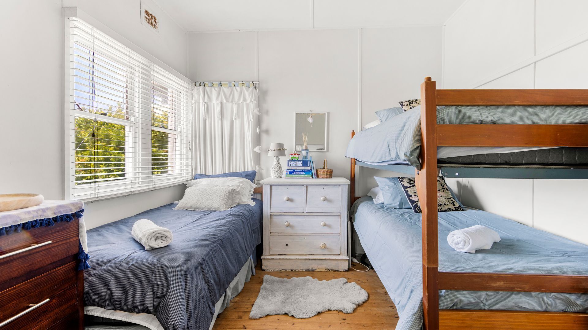 A dorm-style room with bunk beds, a dresser, a desk, and a window with blinds.