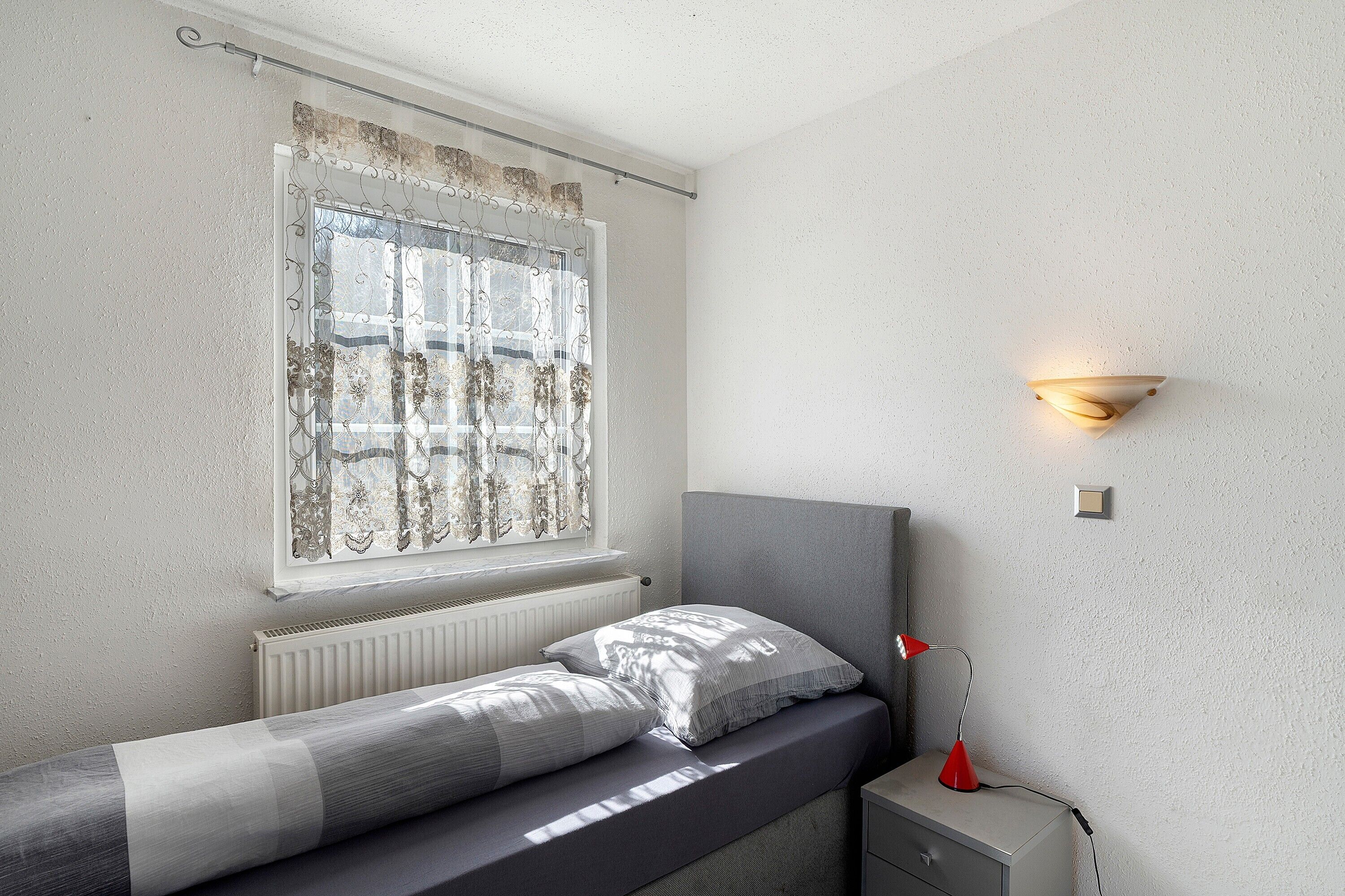 A bedroom with a bed, a window with curtains, a bedside lamp, and a wall-mounted light fixture.