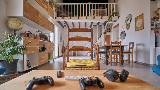 Una sala de estar acogedora con una silla de madera, un reposapiés amarillo y mandos de videojuegos sobre una mesa.