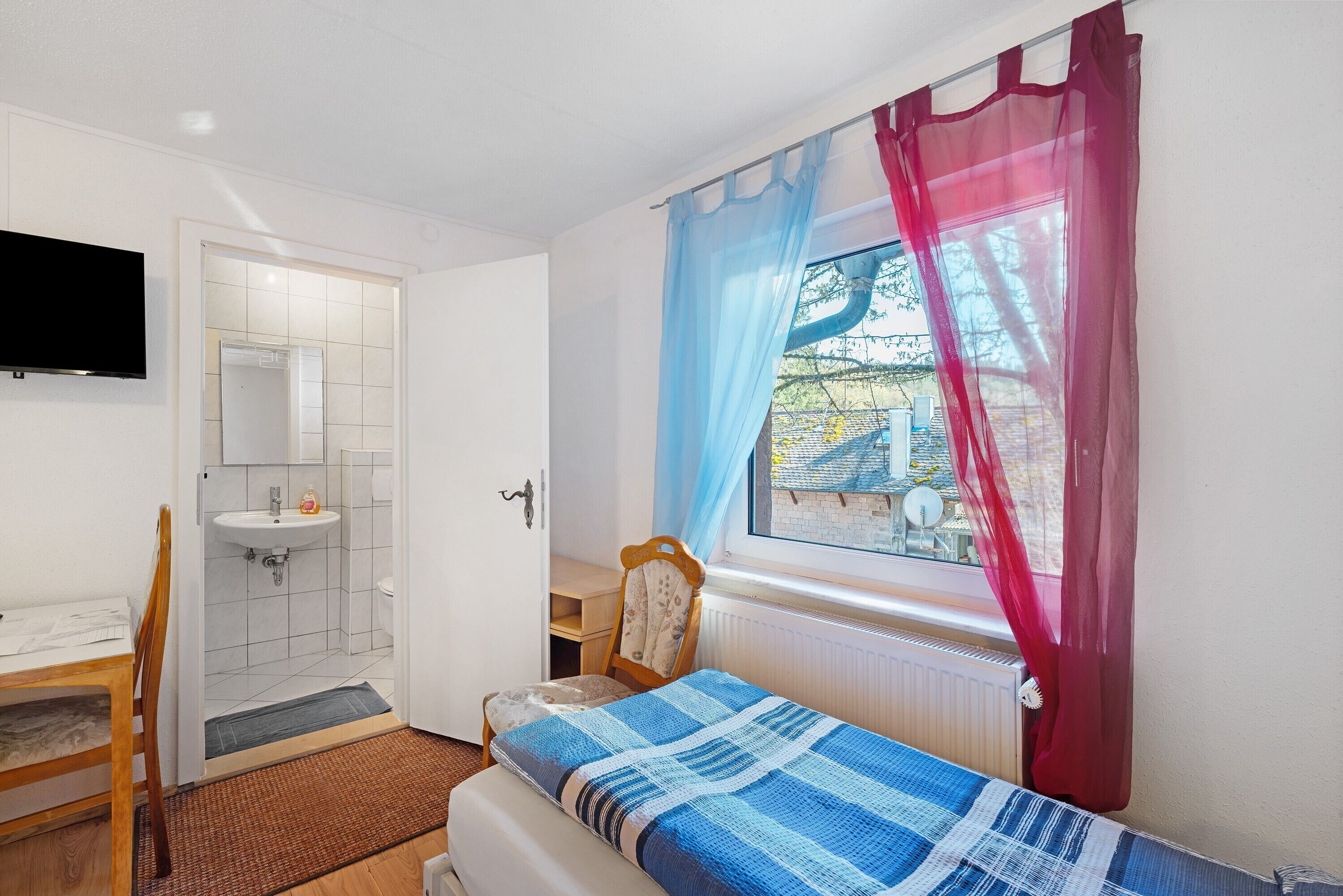 A bedroom with a bed, a chair, a desk, a window with curtains, a TV mounted on the wall, and a bathroom with a sink and mirror.
