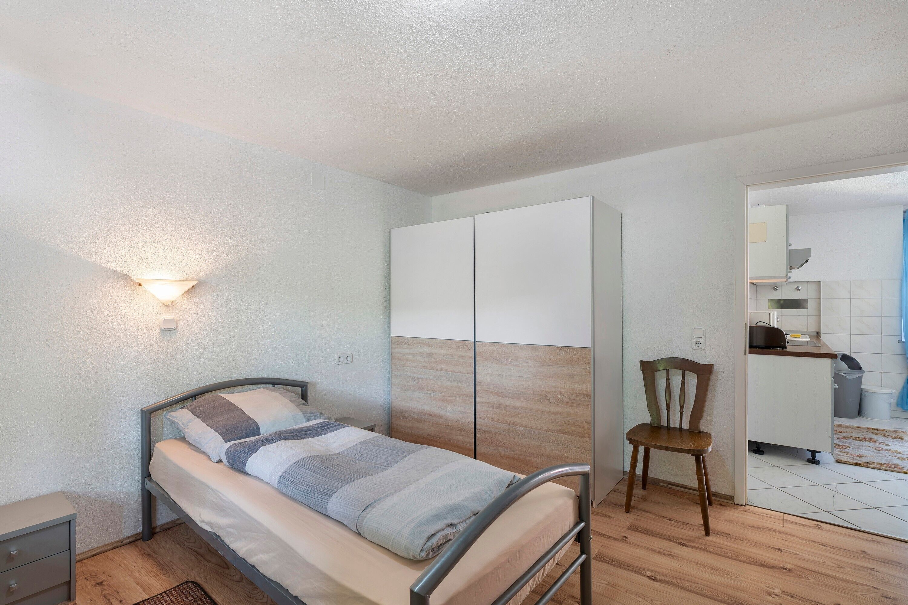 A bedroom with a bed, a chair, a wardrobe, and a kitchenette.