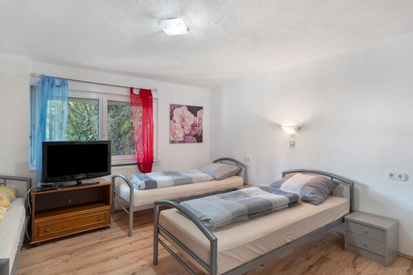 Une chambre avec deux lits simples, un téléviseur posé sur un meuble en bois, une fenêtre avec des rideaux et un luminaire fixé au mur.