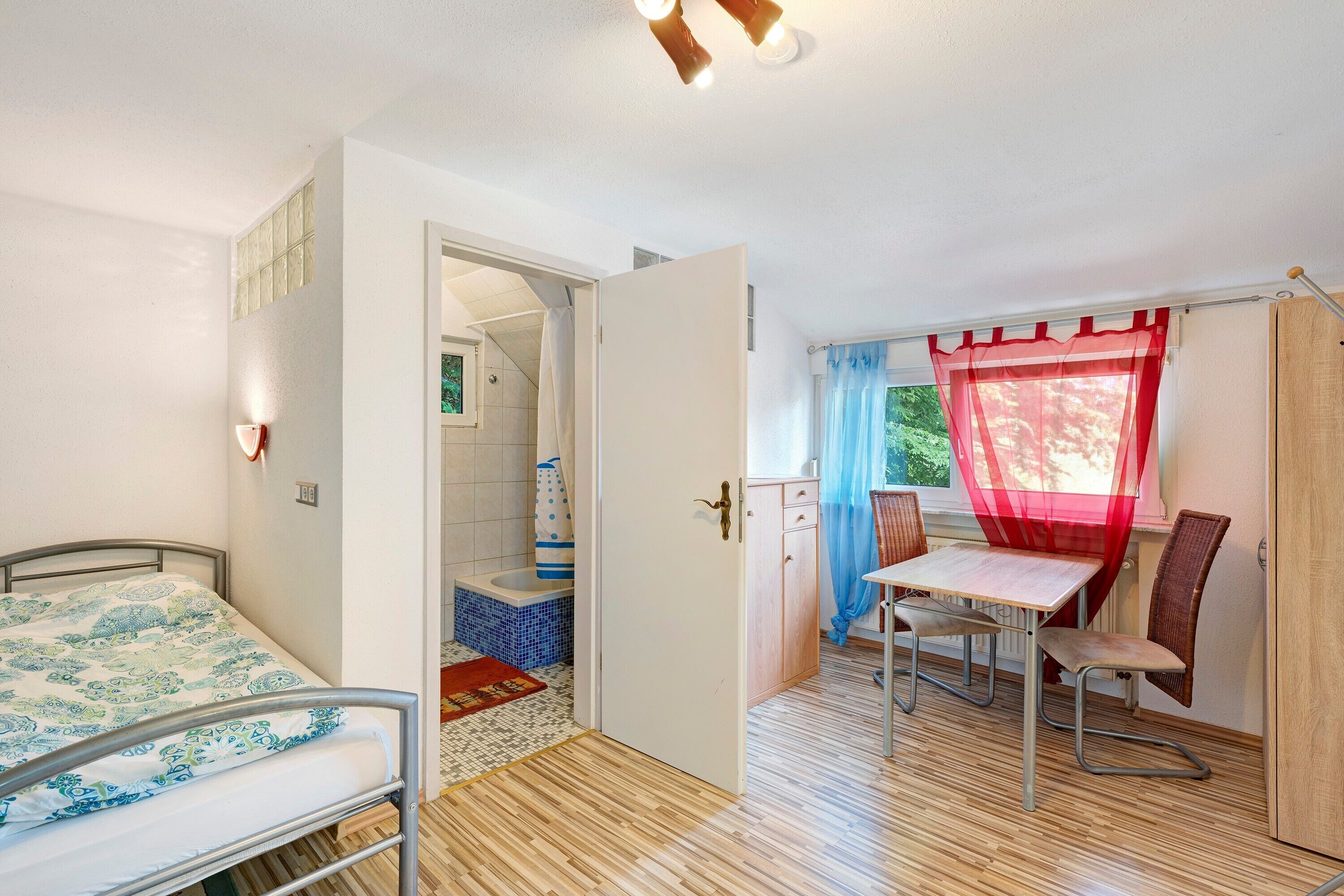 A bedroom with a bed, a bathroom with a shower, a dining area with a table and chairs, and a window with curtains.