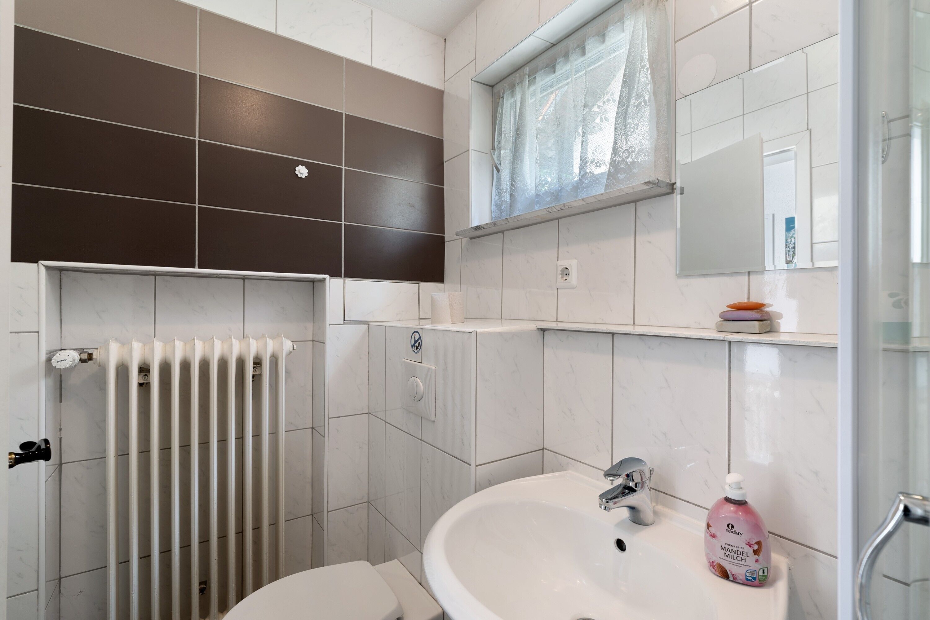 A bathroom with a white sink, a mirror, a window with lace curtains, a radiator, and a toilet.