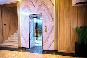 A marble wall with a glass elevator door, a wooden staircase, and a potted plant. - Bourdillon Hotel & Apartments Airport Rd (Abuja)