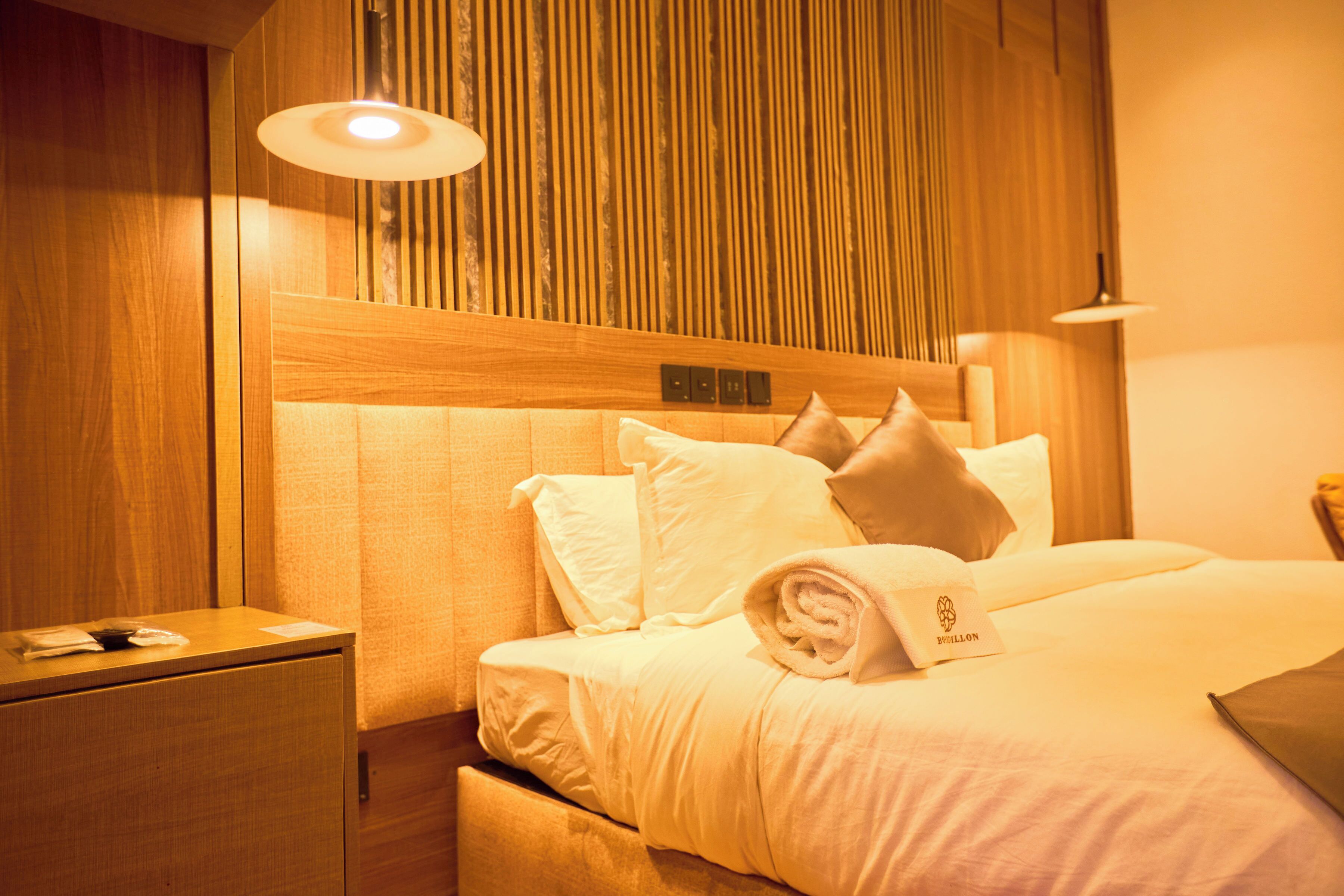 A hotel room with a wooden headboard, a neatly made bed with white linens, a small bedside table, and a wall-mounted light fixture.