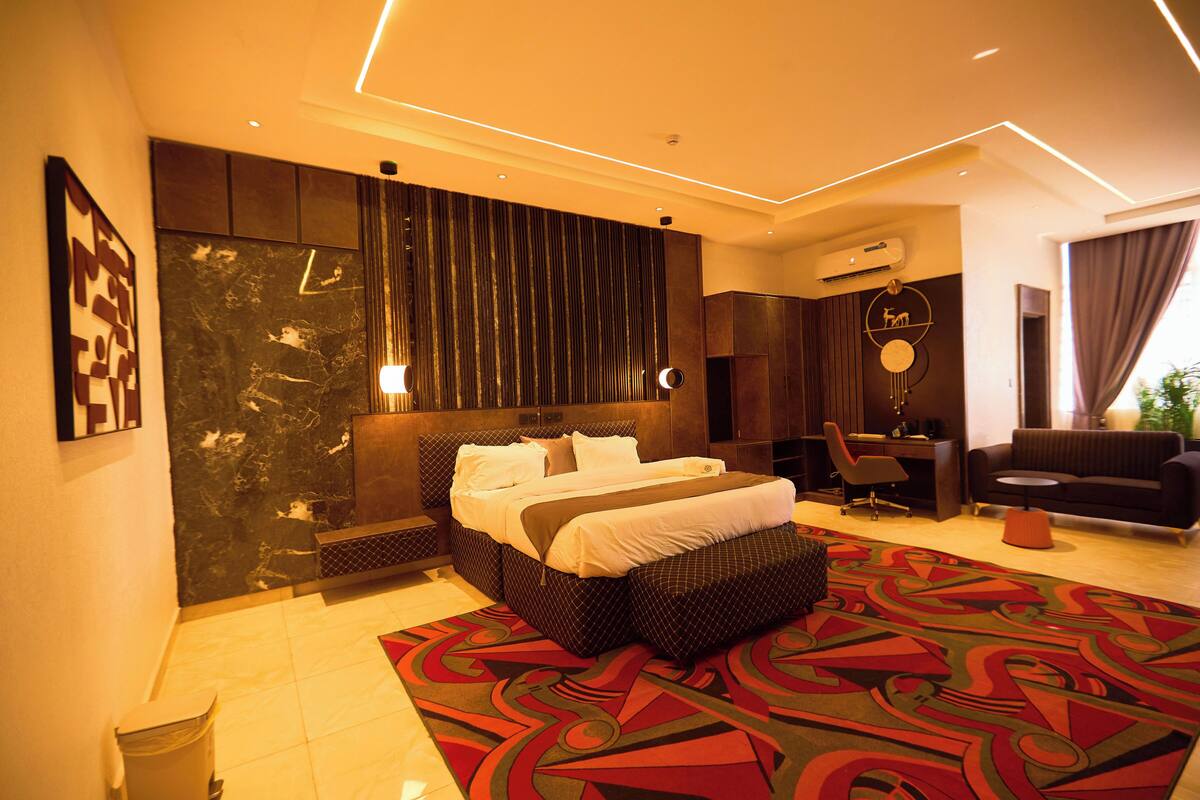 A modern hotel room with a large bed, a desk, a sofa, and a patterned rug.