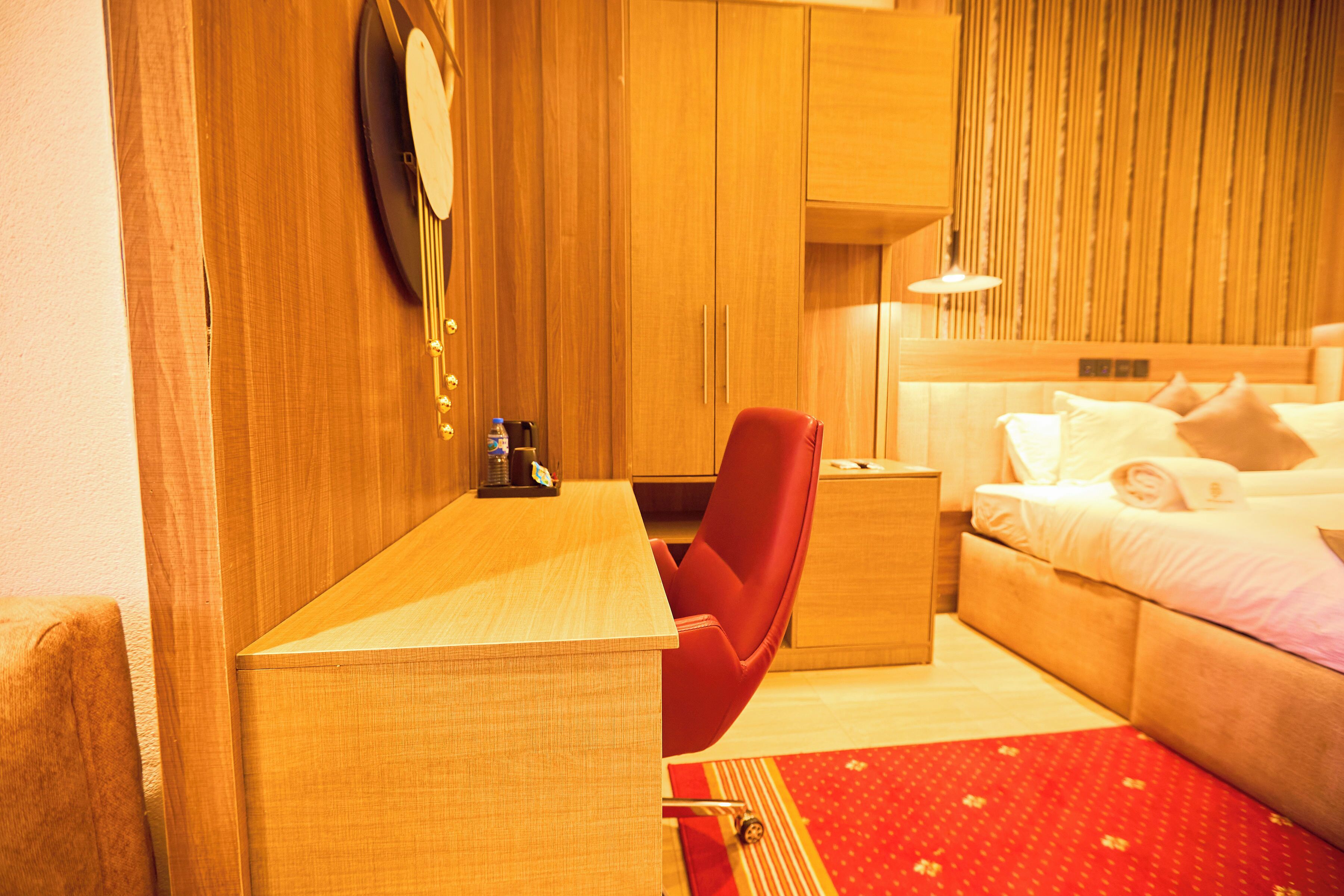 A hotel room with a wooden desk, a red chair, a bed with pillows, and a bedside table.