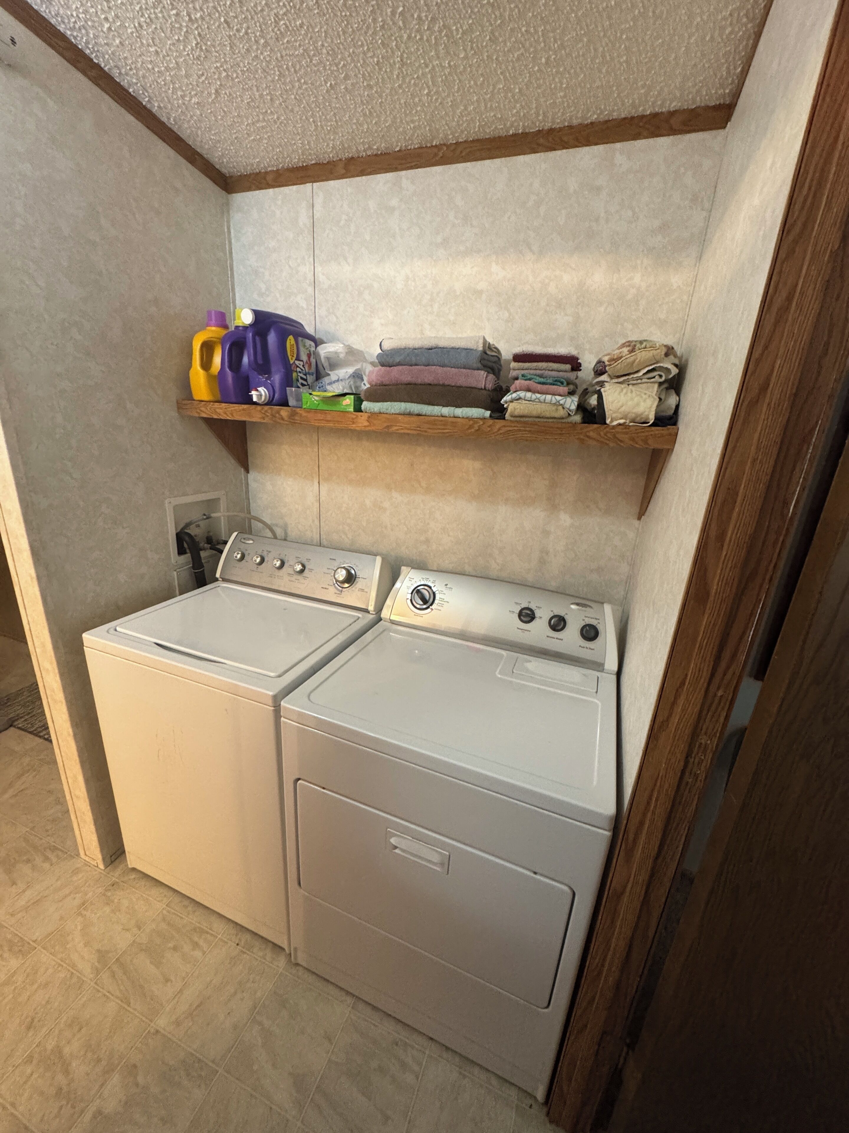 A laundry room with two front-loading washing machines and a shelf above containing folded towels and cleaning supplies.