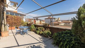 Eine Dachterrasse mit Tisch und Stühlen, Blumentöpfen und Blick auf die umliegenden Gebäude.