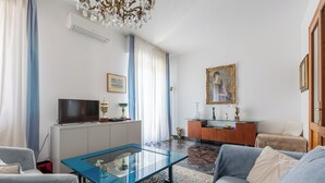 Ein Wohnzimmer mit einem blauen Sofa, einem Glastisch, einem Fernseher, einer Kommode und einem Gemälde an der Wand.