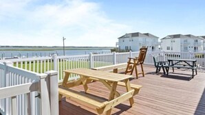 Una terraza de madera con mesas y sillas de picnic, una cerca blanca y edificios al fondo.