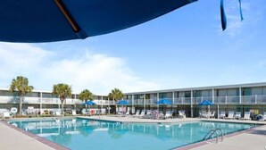 Un área de piscina con sillones de playa y sombrillas, un edificio con balcones y palmeras al fondo.
