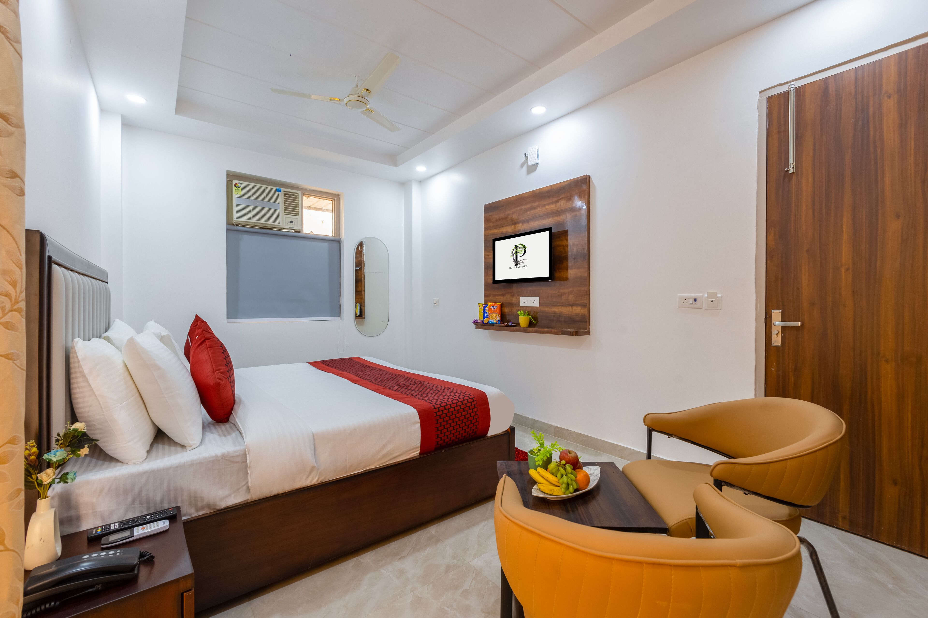 Una camera d'albergo con un letto, una scrivania con una ciotola di frutta, una sedia, una porta in legno e una ventola a soffitto.