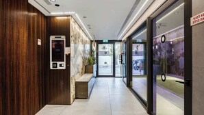 Een moderne hotellobby met een houten wand, een digitaal display voor de lift, een bank en een glazen deur die naar buiten leidt.