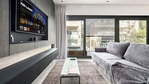 Een moderne woonkamer met een flatscreen televisie aan de muur, een grijze bank, een salontafel en een groot raam met uitzicht op de omgeving.