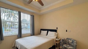 Un dormitorio con cama, mesita de noche, silla, ventana con cortinas y ventilador de techo.