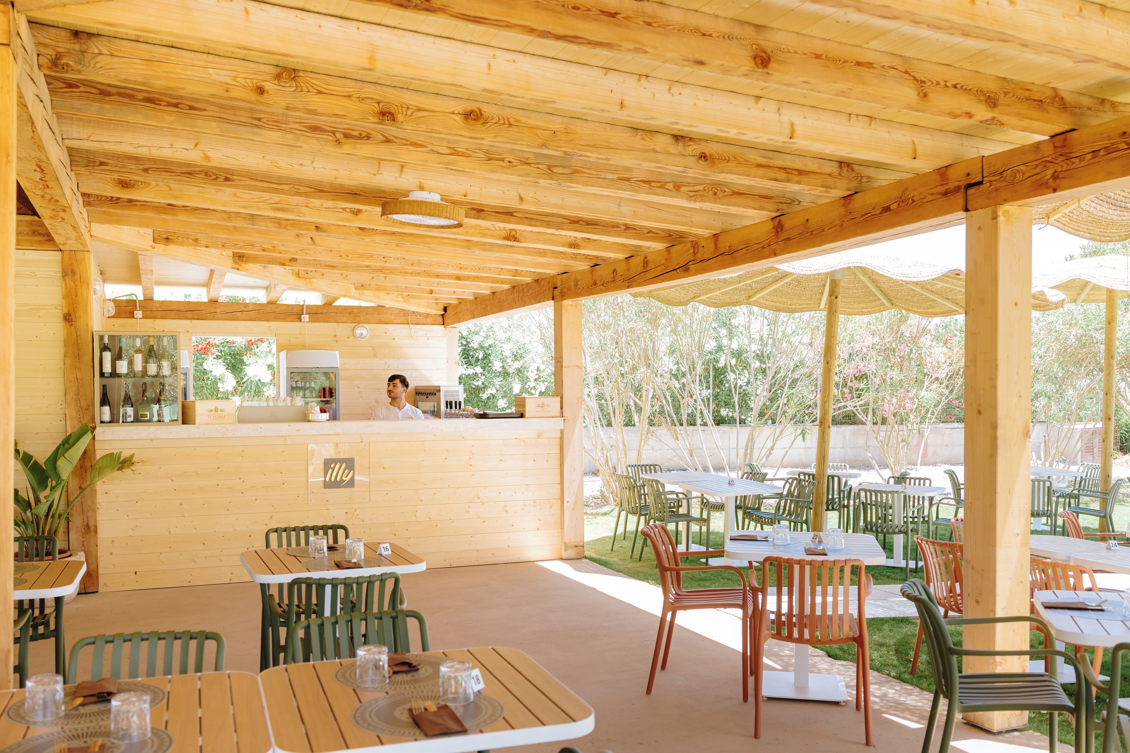 Patio esterno in legno con tavoli e sedie, angolo bar e vista sugli alberi.
