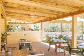 An outdoor wooden patio with tables and chairs, a bar area, and a view of trees. - Valtur Sardegna - Eos Costa Rei  (Costa Rei)