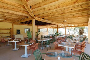 Un espace extérieur aménagé pour les repas, avec des tables et des chaises en bois, un tableau noir servant de menu et des plantes en pot.