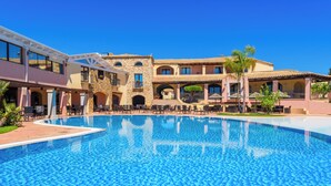 A large outdoor swimming pool with a clear blue water surface, surrounded by a multi-story building with balconies and a patio area. - Valtur Sardegna - Eos Costa Rei  (Costa Rei)