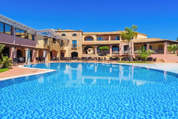 A large outdoor swimming pool with a clear blue water surface, surrounded by a multi-story building with balconies and a patio area. - Valtur Sardegna - Eos Costa Rei  (Costa Rei)
