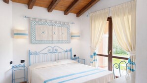 A bedroom with a bed, bedside tables, a wall-mounted lamp, a window with curtains, and a view of the outdoors. - Valtur Sardegna - Eos Costa Rei  (Costa Rei)