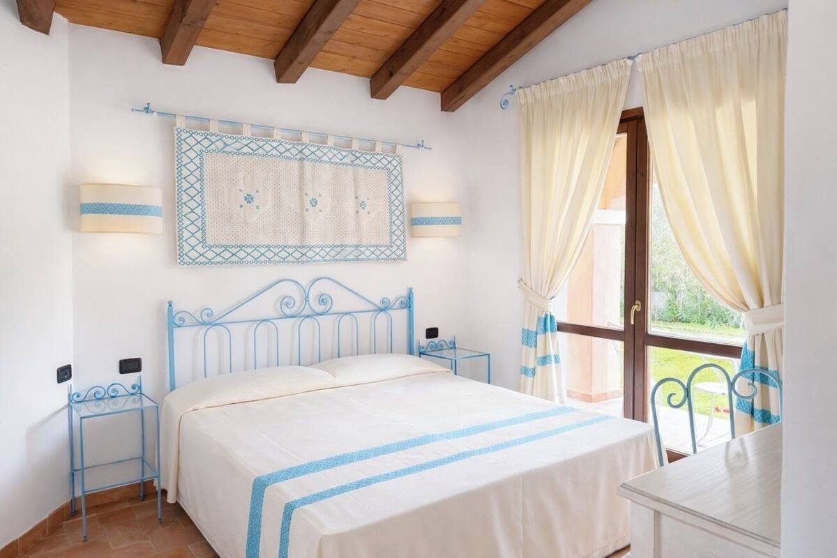 A bedroom with a bed, bedside tables, a wall-mounted lamp, a window with curtains, and a view of the outdoors.