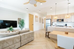 A modern living room with a sofa, TV, and plants. It leads to a kitchen with a fridge, microwave, and dining area.