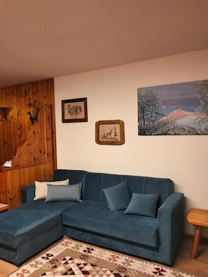 A living room with a blue sofa, a patterned rug, wooden paneling on the left, and two framed pictures on the wall.