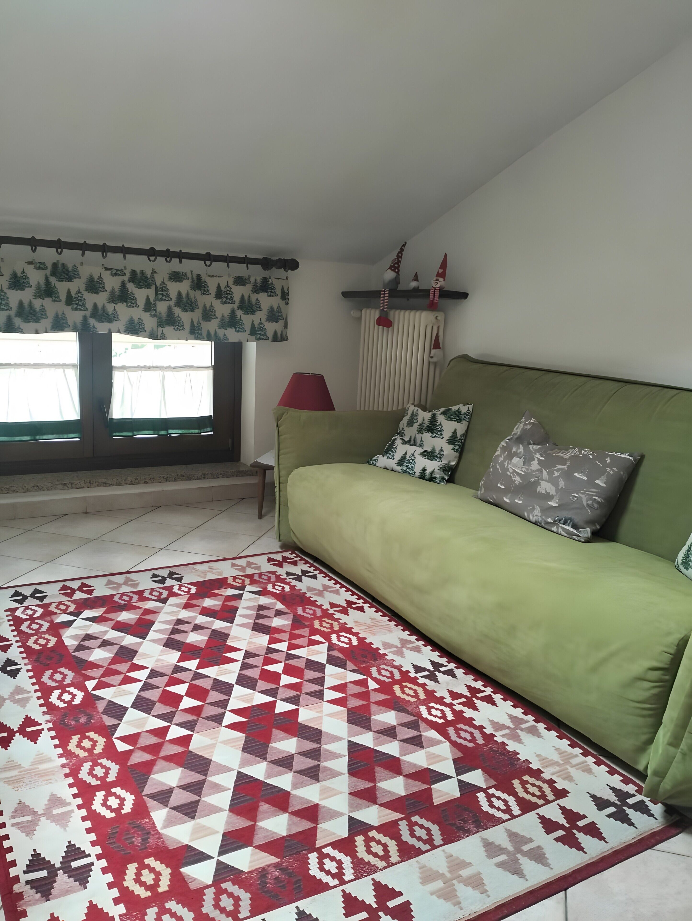 Un sofá verde con cojines estampados, una lámpara roja, una alfombra con diseño y una ventana con cortinas.