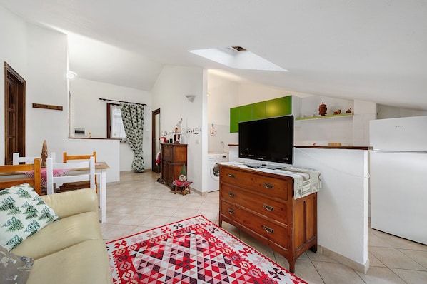 A compact living space with a sofa, a TV, a wooden dresser, a kitchen area with a fridge, and a patterned rug.