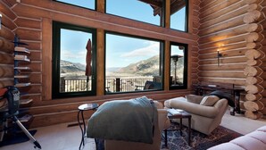 Ein Holzblockhaus-Interieur mit einem großen Fenster, das Blick auf die Berge bietet, eine gemütliche Sitzecke mit einer Couch und Sesseln sowie ein kleiner Tisch.