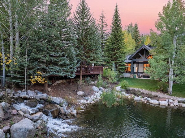 A cabin with a deck, surrounded by trees and a pond.