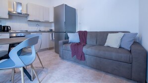 A modern kitchen with a refrigerator, stove, and sink; a grey sofa with cushions; and a dining area with a table and chairs.