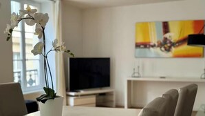 A modern living room with a white orchid plant on a table, a flat-screen TV, and a colorful abstract painting on the wall. - AN EXCEPTIONAL PARISIAN EXPERIENCE CLOSE TO ODEAN & LUXEMBOURG – SAINT GERMAIN (Paris)