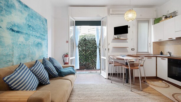 Un salon moderne avec un canapé, des coussins à rayures, un grand tableau abstrait et une vue sur un balcon, visible à travers une porte vitrée.