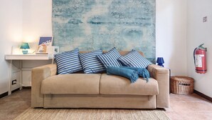 Un canapé beige agrémenté de coussins à rayures, une couverture bleue, un tableau abstrait accroché au mur, une petite table d’appoint avec une lampe et un extincteur.