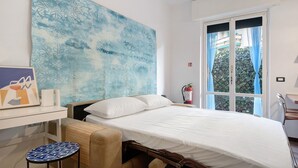 Une chambre à coucher comprenant un lit, un bureau, une chaise, une fenêtre avec des rideaux et un mur orné d’un tableau abstrait bleu.
